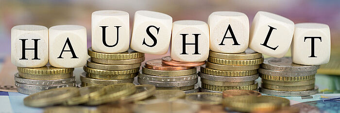 Foto mit Geldmünzen und Würfeln mit Buchstaben. Auf den Buchstaben steht das Wort "Haushalt" geschrieben.